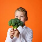 A cheerful young boy holds fresh broccoli, promoting healthy eating against a vibrant orange backdrop.